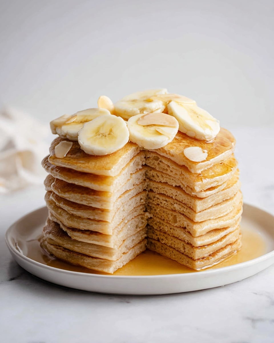 A tall stack of 12 light golden brown pancakes sits on a round white plate, with a wedge cut out to show the even, smooth layers inside. The pancakes are soft and thin, with a slight shine from syrup that glistens on top and drips down the sides. On the top, there are about five slices of pale yellow banana arranged neatly, with a few small almond slivers scattered among them. The scene is set against a white marbled surface and a clean, soft white background. Photo taken with an iphone --ar 4:5 --v 7