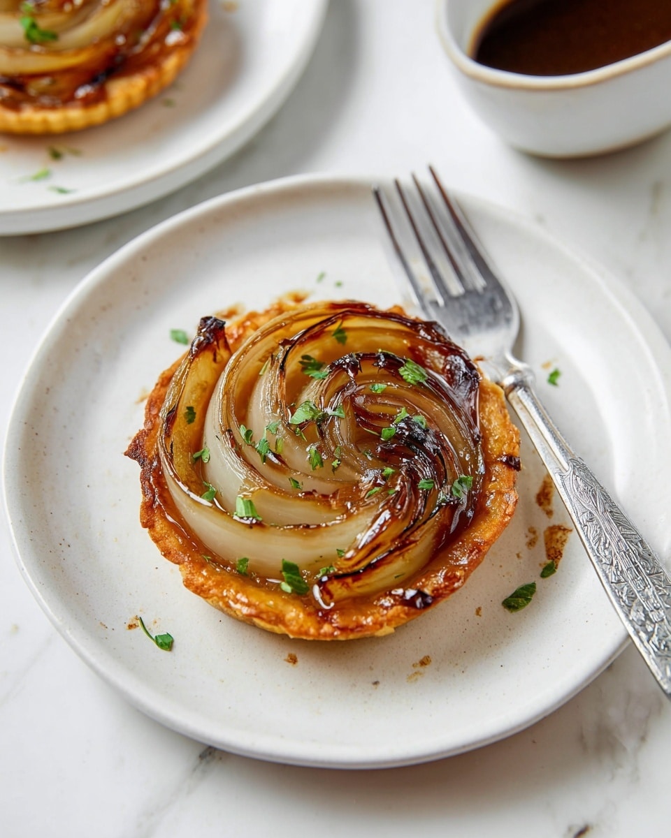 A small round tart sits in the center of a white plate with a slightly raised edge, placed on a white marbled surface. The tart has a golden-brown, crispy crust as the base layer. On top, there are several thick onion slices arranged in a flower shape, showing a mix of translucent white and lightly charred brown tones. The onions have a shiny glaze that reflects light, making them look soft and juicy. Scattered evenly across the top are small pieces of fresh green herbs. A silver fork with a simple etched handle rests on the plate's right side. In the background, there is another plate with a similar tart and a small white bowl holding a thick dark brown sauce. Photo taken with an iphone --ar 4:5 --v 7