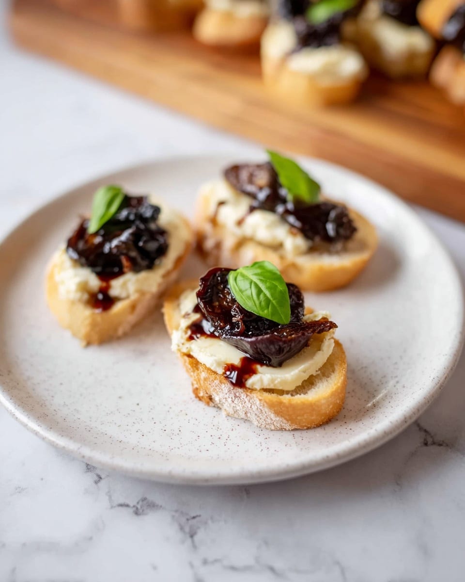 Three small toasted baguette slices are placed on a white plate with light speckles. Each slice has a creamy, pale yellow layer of soft cheese spread evenly on top. On the cheese, there are pieces of dark brown, glossy dried fruit, possibly figs or prunes, adding a rich contrast. A few thin trails of dark red balsamic glaze are drizzled over the cheese and fruit layers. Each piece is finished with a fresh green basil leaf on top, adding a bright splash of color. The plate is set on a white marbled surface with a wooden board blurred in the background holding more of these bite-sized snacks. Photo taken with an iphone --ar 4:5 --v 7