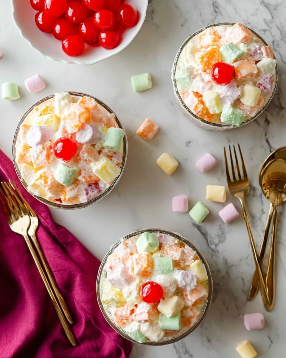 The image shows three glass bowls filled with a creamy, colorful fruit salad placed on a white marbled surface. Each bowl contains a mix of soft white cream and small chunks of pastel-colored marshmallows in light green, yellow, pink, and orange, along with bright orange fruit pieces. One bowl is topped with a shiny red cherry, and extra small pastel marshmallows are scattered around the bowls on the surface. There is a small white dish filled with shiny red cherries in the upper left corner. Two golden forks are placed on the surface, with a rich magenta cloth napkin also visible near the bottom left corner. Photo taken with an iphone --ar 4:5 --v 7