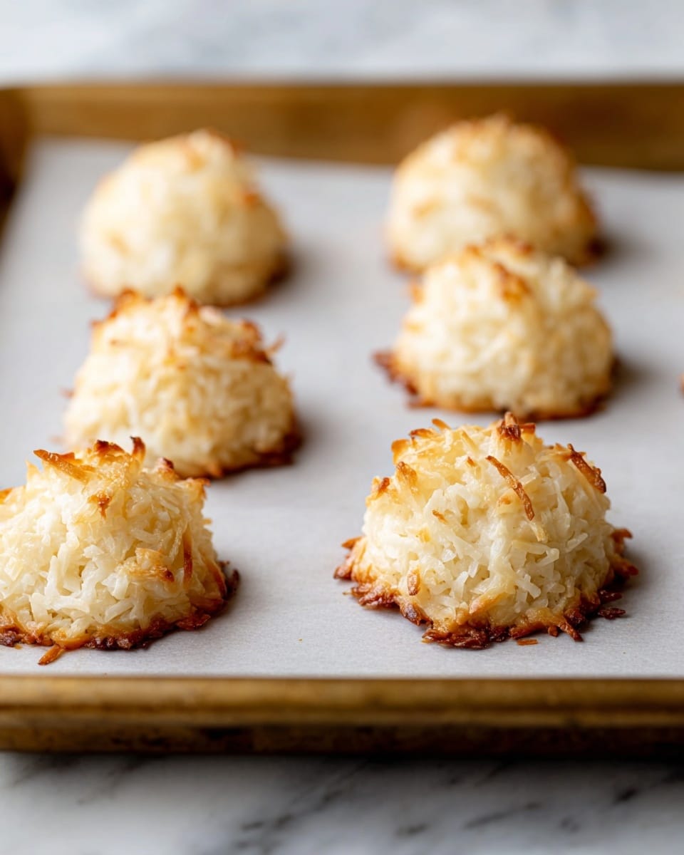 There are six light golden brown coconut macaroons on a baking sheet lined with white paper. Each macaroon is shaped like a small mound, with shredded coconut on the surface giving a rough and textured look. The edges are a little darker and crispier, while the tops are soft and pale. The baking sheet has a brass color, and the background is a soft white marbled texture. photo taken with an iphone --ar 4:5 --v 7
