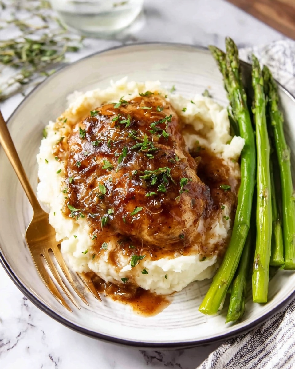 A white plate with a thin black rim holds a meal layered with creamy white mashed potatoes as the base, topped by a brown cooked chicken piece covered in a rich, glossy sauce sprinkled with finely chopped green herbs. To the right of the chicken, there is a neat bundle of bright green asparagus stalks, slightly shiny from cooking. A gold fork rests on the left edge of the plate on a white marbled surface photo taken with an iphone --ar 4:5 --v 7