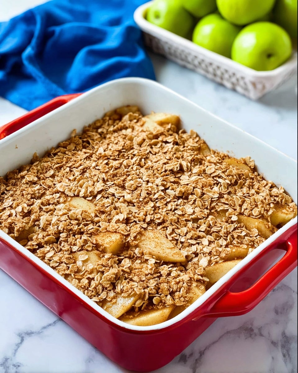 A square white baking dish with red handles holds a baked apple crisp dessert. The bottom layer consists of soft, golden-brown cooked apple slices, and the top layer is a rough, textured oat crumble with a light brown color, evenly spread across the apples. The dish sits on a white marbled surface with a blue cloth and a white basket of green apples in the background. Photo taken with an iphone --ar 4:5 --v 7