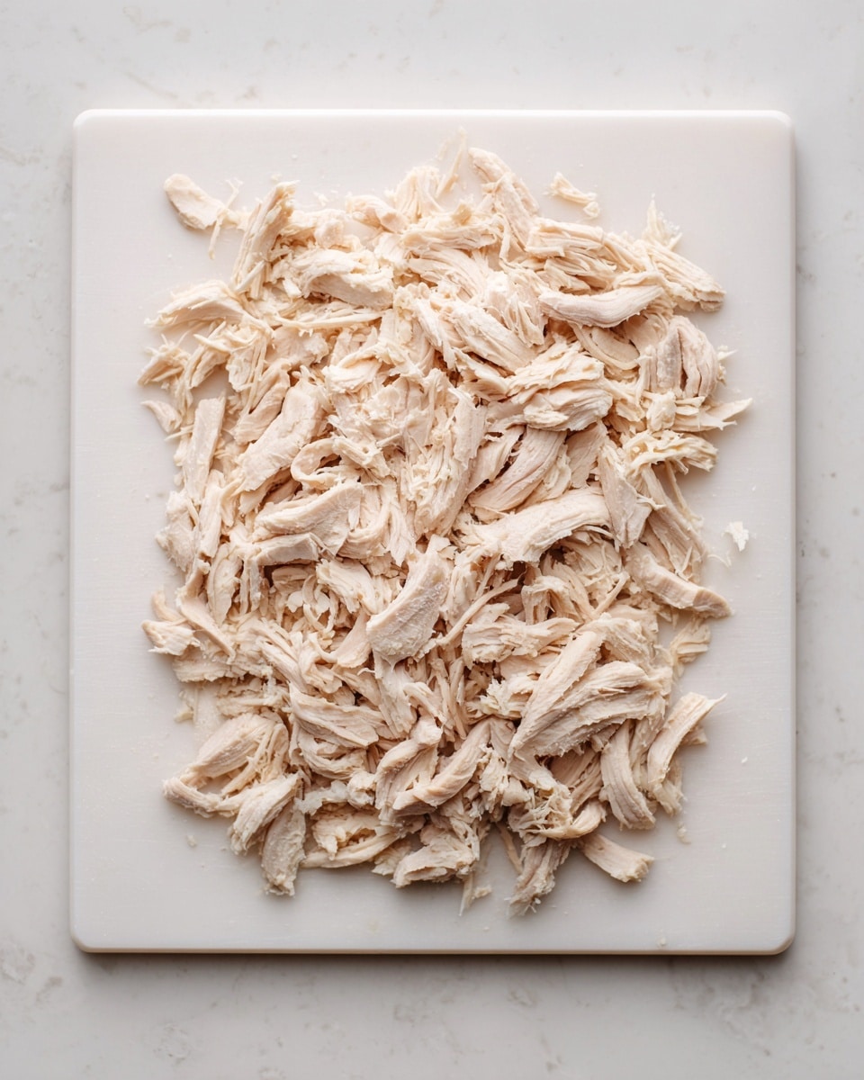 The image shows a white cutting board placed on a white marbled surface. On the cutting board, there is one layer of shredded light beige chicken meat spread out evenly. The chicken pieces have a fibrous texture, with visible strands and slight variation in color from light beige to a slightly darker shade. The whole scene looks clean and simple with no other objects or layers present. Photo taken with an iphone --ar 4:5 --v 7