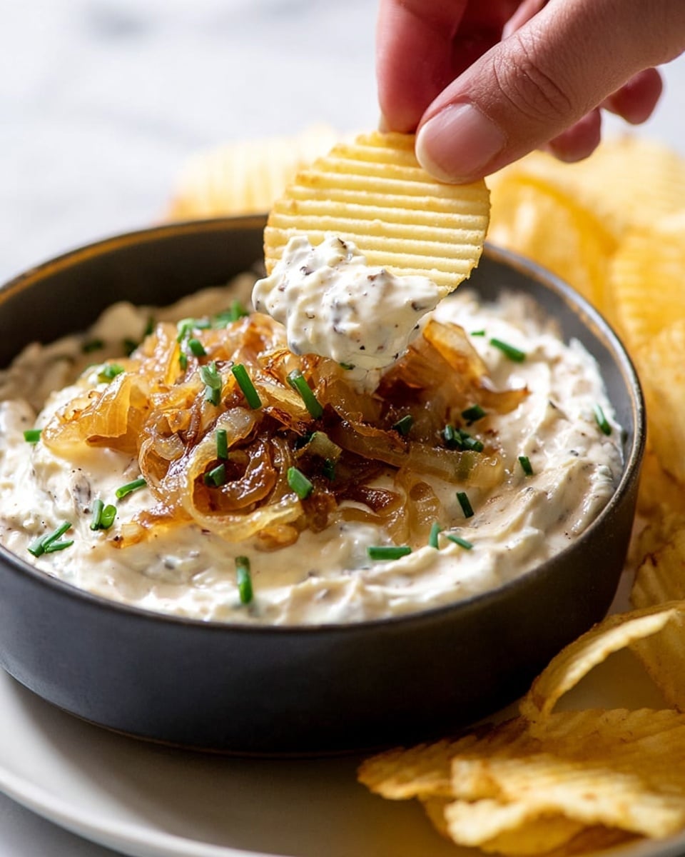 A close-up image shows a woman's hand dipping a light yellow ridged potato chip into a creamy dip inside a dark bowl. The dip is white with small bits of black pepper and has a thick, smooth texture. On top of the dip is a layer of caramelized onions that are golden brown and slightly shiny, sprinkled with small pieces of fresh green chives. The bowl sits on a white plate with more ridged potato chips around it, all placed on a white marbled surface. photo taken with an iphone --ar 4:5 --v 7