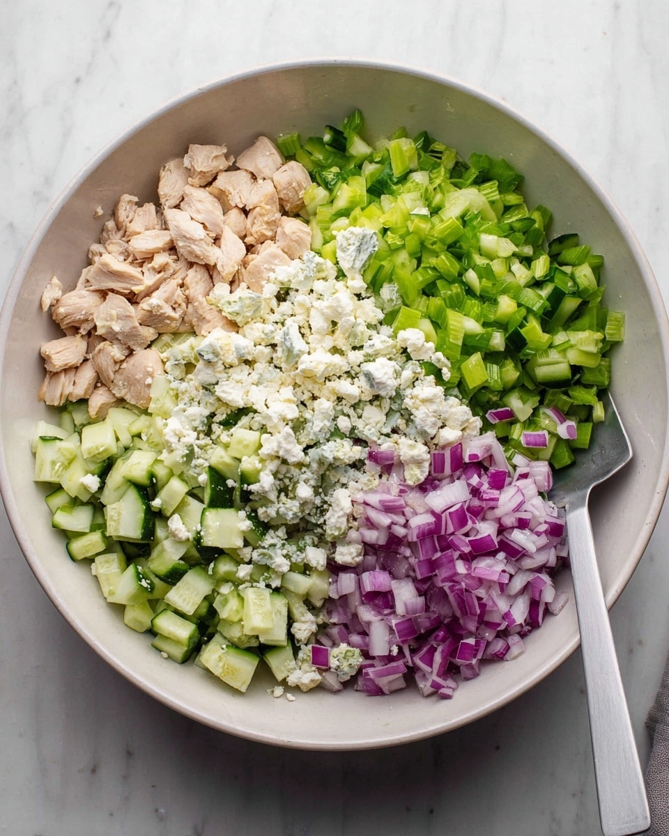 A large white bowl sits on a white marbled surface filled with five distinct layers of ingredients arranged side by side: cooked, light tan chicken pieces on the left, bright green chopped celery next to the chicken, chopped cucumber pieces with a darker green skin on the right, crumbled white cheese piled over part of the cucumber, and finely diced purple-red onions at the bottom near the celery. A light gray spatula rests to the right of the bowl. The lighting is soft and natural, highlighting the fresh textures. photo taken with an iphone --ar 4:5 --v 7
