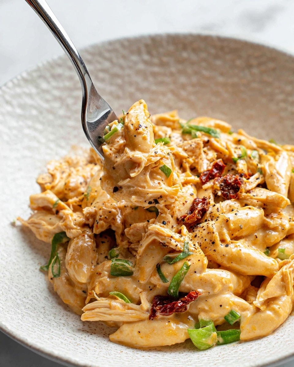 The dish shows a creamy pasta served on a white textured plate over a white marbled surface. The pasta is short and oval-shaped, covered in a thick, light orange sauce. Mixed in are pieces of shredded chicken, some small bright green vegetable pieces, and bits of sun-dried tomatoes adding red accents. A fork lifts a portion, showing the creamy texture coating each ingredient with some black pepper sprinkled on top. The overall look is rich, warm, and inviting. Photo taken with an iphone --ar 4:5 --v 7