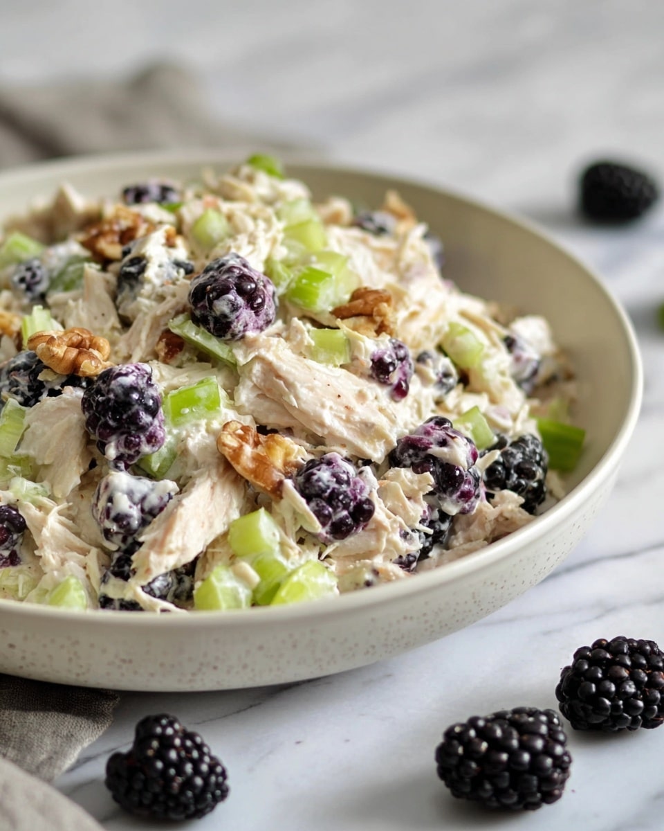 The image shows a bowl filled with a creamy salad that has several layers and colors. The main layer is shredded light beige chicken mixed with small pieces of bright green celery. Scattered through the salad are dark purple-black blackberries that add a deep color contrast. There are small chunks of light brown walnuts mixed in, adding texture. The creamy white sauce covers all ingredients evenly. The bowl is a white ceramic with a smooth finish, sitting on a white marbled surface. In the foreground, there are a few whole blackberries placed casually. The photo is clear and natural, showing close details of the salad, photo taken with an iphone --ar 4:5 --v 7