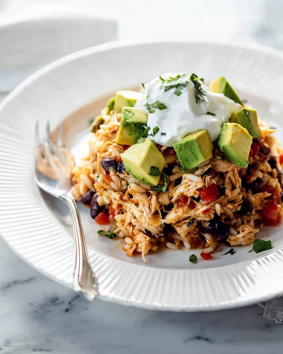 A white plate with ridged edges holds a mound of mixed ingredients. The bottom layer is light brown shredded chicken mixed with small black beans, pieces of white rice, red bell pepper bits, and chopped green herbs scattered throughout. Cubed green avocado pieces are placed on top and around the mix, adding pops of bright green. Sitting on the very top center is a dollop of smooth white sour cream. A silver fork rests on the left side of the plate on a white marbled surface in soft natural light. photo taken with an iphone --ar 4:5 --v 7