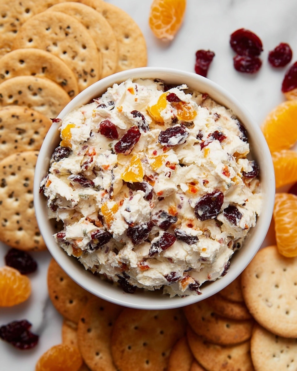A close-up view of a round, golden-brown cracker held by a woman's hand, showing a scoop of creamy white cheese mixed with bright orange chunks and dark red dried fruit pieces. The textures are soft and crumbly in the cheese mixture, contrasting with the smooth and slightly crisp cracker. The background is softly blurred with hints of more crackers, placed on a white marbled surface. photo taken with an iphone --ar 4:5 --v 7