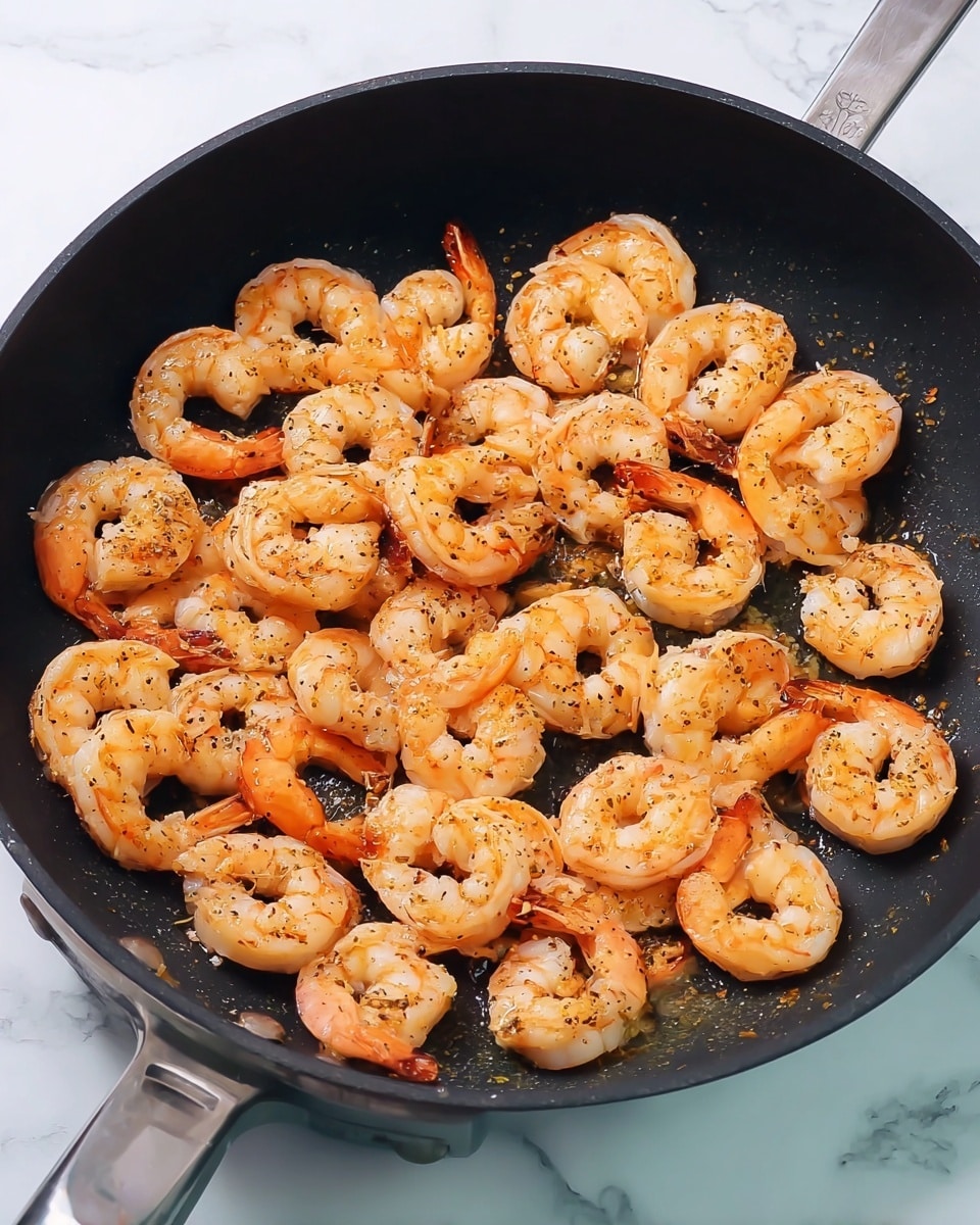 A black cooking pan filled with a single layer of cooked shrimp, each shrimp curled and pink with a light orange tint, speckled with black pepper and light seasoning, arranged closely but not overlapping. The pan sits on a white marbled surface, and the metal handle of the pan is visible on the right side. Photo taken with an iphone --ar 4:5 --v 7