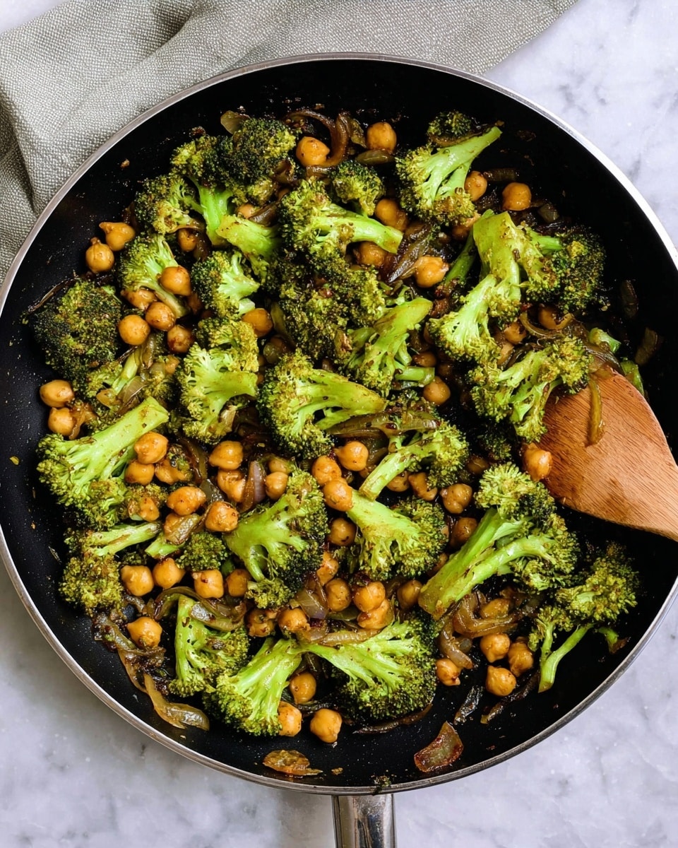 The image shows a black frying pan filled with cooked broccoli and chickpeas mixed with small pieces of onion. The broccoli is bright green with a slightly browned, soft texture, and is cut into medium-sized florets spread evenly all over the pan. The chickpeas are light brown, smoothly round, and scattered throughout, mingling with the onions which are soft and golden. A wooden spatula rests inside the pan on the right side. The pan is placed on a white marbled surface with a light gray cloth partially visible in the top left corner. Photo taken with an iphone --ar 4:5 --v 7