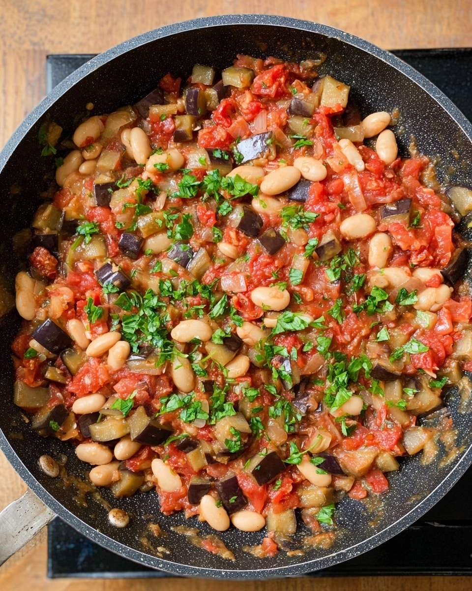 The image shows a black frying pan filled with a cooked dish of chopped eggplants, white beans, and diced tomatoes mixed together. The colors are mostly light brown and red from the vegetables with white beans scattered throughout. Bright green chopped herbs are sprinkled on top, adding a fresh look. The pan rests on a black stovetop, and the background is a warm wooden surface. The food looks soft with a chunky texture from the beans and vegetables, and the herbs add a touch of brightness. photo taken with an iphone --ar 4:5 --v 7