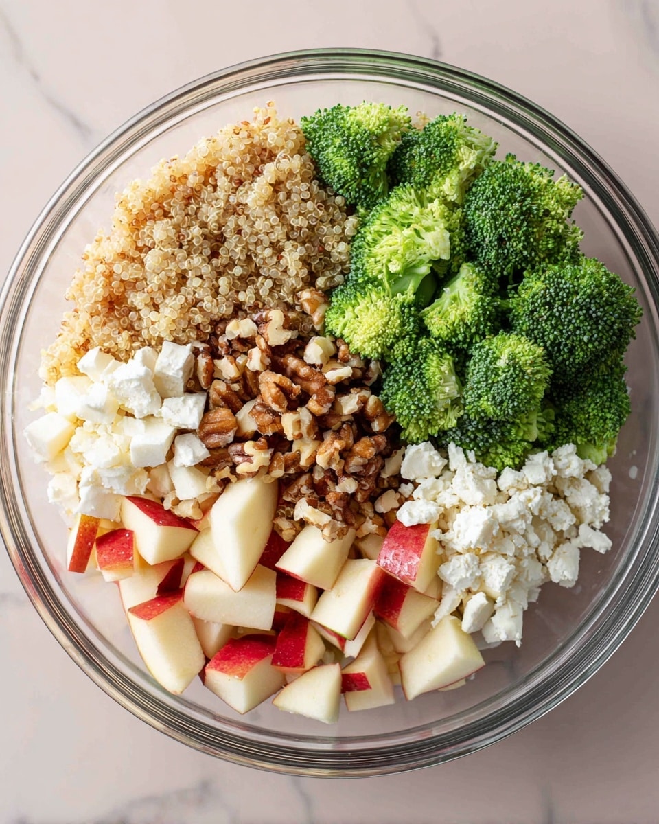 A clear glass bowl filled with five layers arranged side by side: bright green broccoli florets at the top right, white crumbly cheese just below the broccoli, small cubed red and white apples at the lower right, a mix of chopped brown nuts in the middle bottom, and light tan cooked quinoa filling the left side. The bowl sits on a white marbled surface. photo taken with an iphone --ar 4:5 --v 7