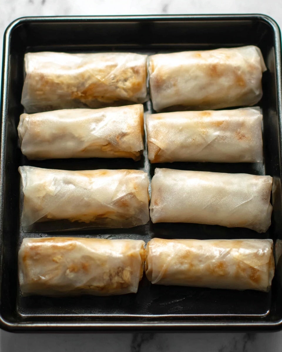 Six rolled spring rolls are placed in two rows on a shiny black baking tray. Each roll is wrapped in translucent rice paper that shows the fillings inside, which are light brown and beige with some small bits of darker color. The rolls look moist and soft with a gentle sheen on the surface. The black tray contrasts with the light colors of the rolls, making them stand out. The background is a white marbled texture. Photo taken with an iphone --ar 4:5 --v 7