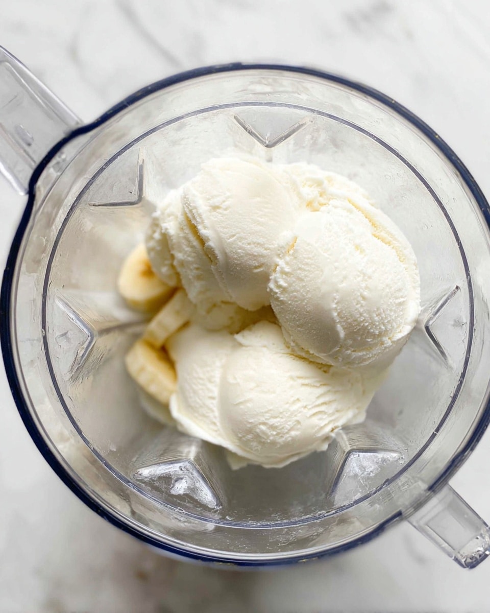 Inside a clear blender jar, there are three scoops of white ice cream with a smooth and creamy texture, resting closely together. Underneath one side of the ice cream, there are small slices of light yellow banana pieces barely visible. The blender jar is set on a white marbled surface. The lighting is bright and natural, highlighting the softness and coolness of the ice cream and the slight shine of the blender's plastic walls photo taken with an iphone --ar 4:5 --v 7