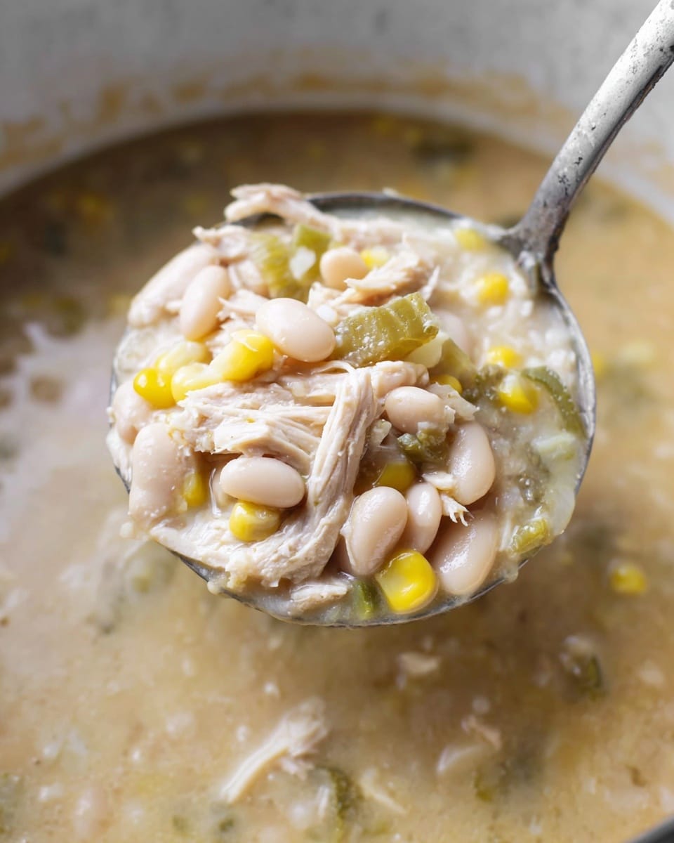 A close-up of a ladle full of thick soup showing three main layers: the base layer is creamy beige broth with small white beans mixed in, the middle layer consists of tender pale shredded chicken pieces, and the top layer includes whole white beans, bright yellow corn kernels, and small green vegetable bits. The ladle is silver and holds a dense mix of the ingredients, with the creamy broth surrounding it inside a pot. The background is a soft white marbled surface. Photo taken with an iphone --ar 4:5 --v 7