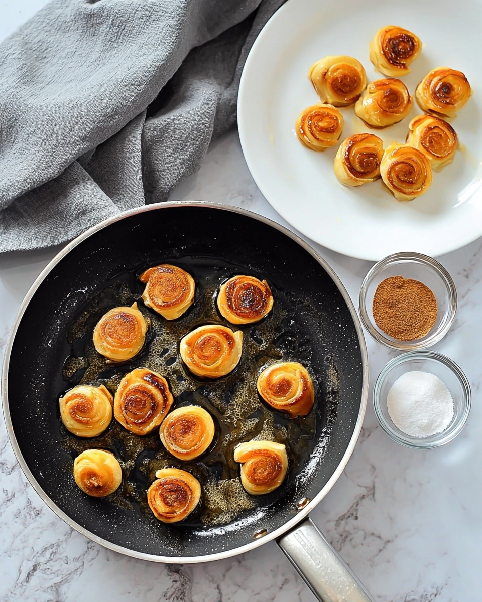 A black frying pan holds about thirteen small, golden-brown spiral rolls frying in melted butter, with crispy edges and soft centers, spread evenly inside. Nearby, a white plate displays more of these golden pastry spirals arranged closely together. Two small clear glass bowls sit on a white marbled surface below the pan; one with white sugar and the other with brownish cinnamon powder. A grey cloth is draped in the upper left corner of the white marbled background. photo taken with an iphone --ar 4:5 --v 7