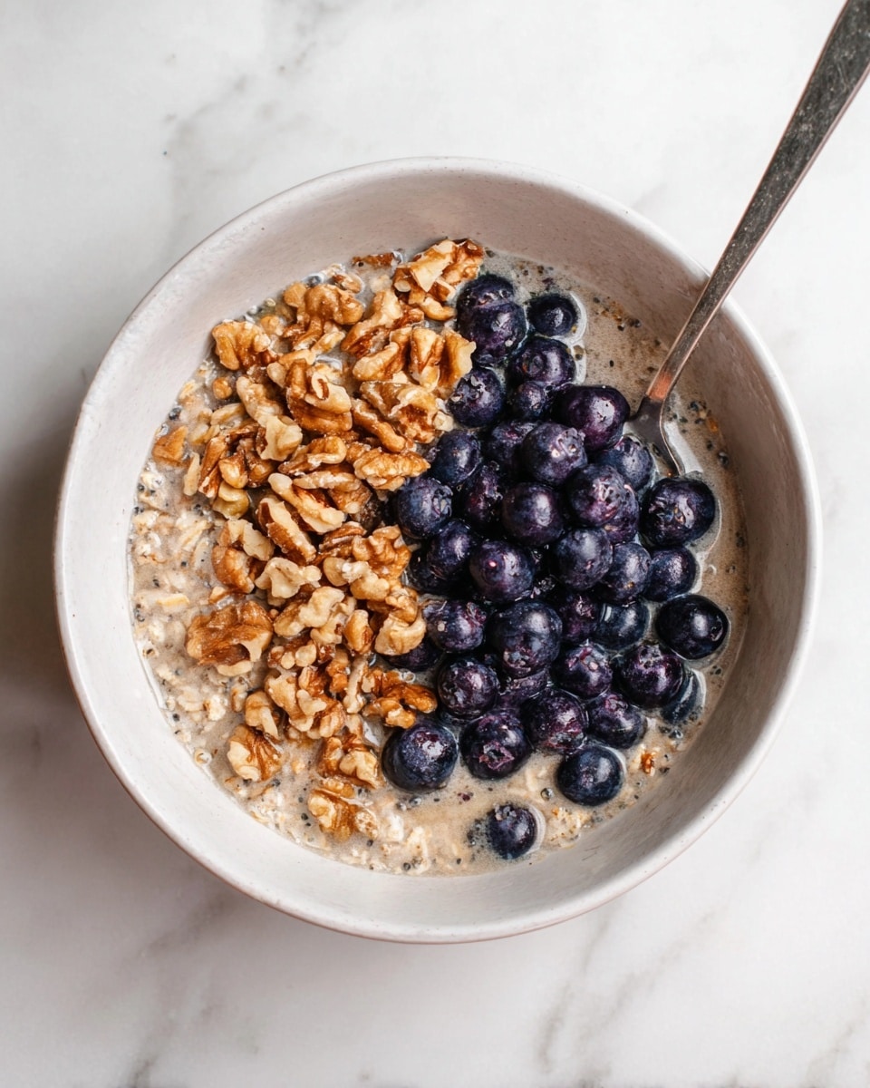 A white bowl filled with soaked oats in a light brown milky liquid, topped with two main layers: on the top left half, a heap of broken walnut pieces showing rough texture and warm brown color; on the bottom right half, a cluster of shiny, fresh blueberries with deep blue and purple hues. A silver spoon rests inside the bowl on the right side, partially submerged in the oats. The bowl sits on a white marbled surface. photo taken with an iphone --ar 4:5 --v 7