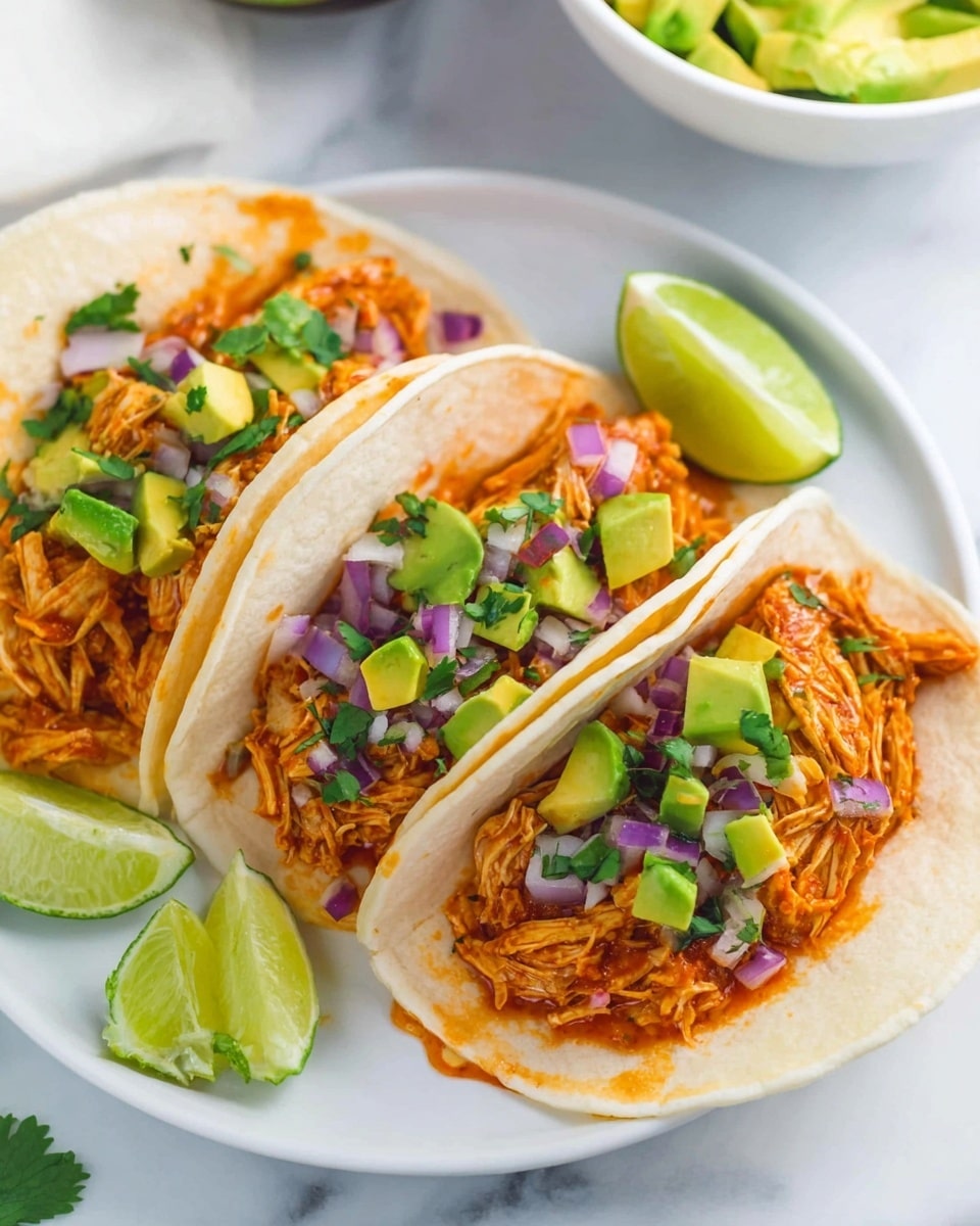 Three soft white tortillas are folded in a white plate, each filled with shredded orange chicken coated in sauce. Topping the chicken are small cubes of green avocado and bits of purple red onion, with green cilantro leaves scattered among them. Around the edges of the plate, there are three lime wedges placed for garnish. In the background, there is a white marbled surface and a white bowl with more avocado pieces visible. Photo taken with an iphone --ar 4:5 --v 7