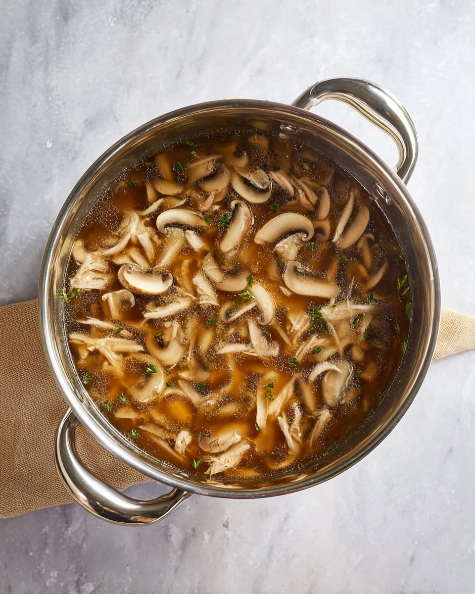 A shiny silver pot filled with a brown broth contains many thinly sliced light brown mushrooms and small pieces of white shredded meat, evenly mixed throughout the soup. The pot has two curved handles on each side and sits on a white marbled surface with a beige cloth partially visible nearby. The broth looks clear but richly flavored with bits of herbs scattered inside. photo taken with an iphone --ar 4:5 --v 7
