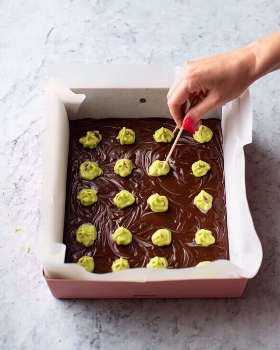 A white baking tray lined with parchment paper filled with a thick, dark brown chocolate batter. On top of the batter, there are even, small dollops of light green batter spread out in rows. A woman's hand with red nail polish is using a toothpick to swirl the two batters together, creating a marbled effect. The baking tray is resting on a surface with a white marbled texture. Photo taken with an iphone --ar 4:5 --v 7