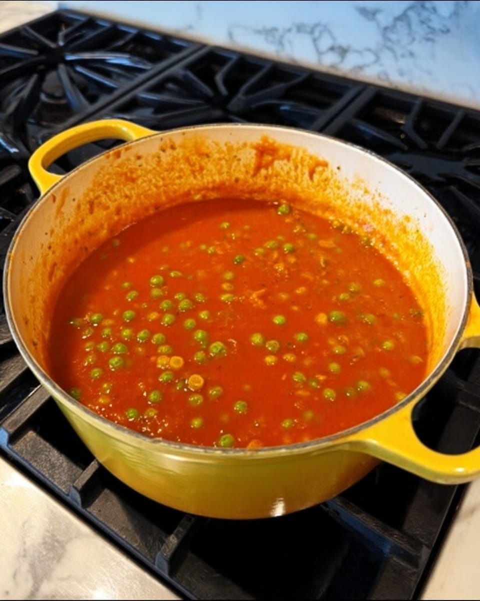 A yellow pot with two handles holds a thick orange-red tomato sauce mixed with bright green peas. The sauce looks smooth with some small bits of tomato and spices visible. The pot sits on a black stove with a white marbled surface underneath. The inside rim of the pot has some sauce stains, showing it is being cooked. photo taken with an iphone --ar 4:5 --v 7