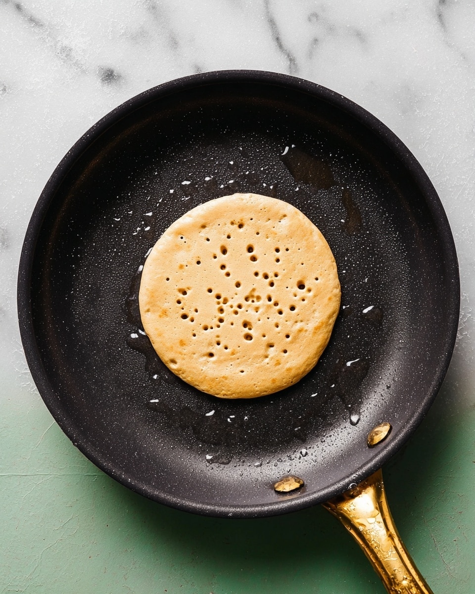 A single round pancake is cooking in the center of a black pan with a golden handle. The pancake is light golden brown with small bubbles and holes forming on the surface, showing it is in the process of cooking. The pan has small droplets of oil giving a slight shine, and the background shows a white marbled surface. The photo taken with an iphone --ar 4:5 --v 7