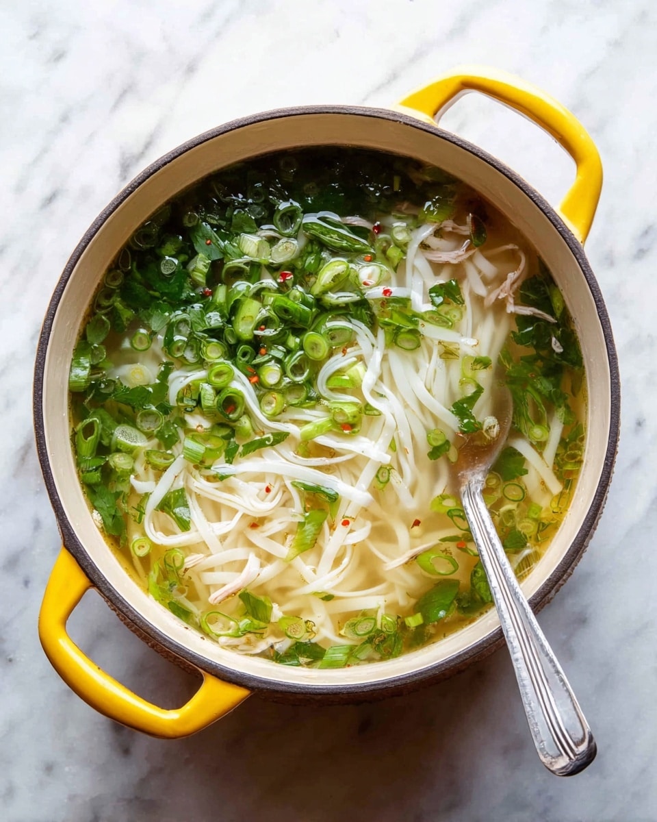 A yellow pot filled with clear broth soup has a layer of flat white noodles resting gently in the middle and bottom. On top, there is a thick cover of bright green chopped spring onions and leafy herbs scattered evenly over the soup surface. There are thin slices of some light-colored meat partially visible under the broth near the noodles. A silver spoon is placed on the right side inside the pot. The pot sits on a white marbled surface with soft natural lighting. Photo taken with an iphone --ar 4:5 --v 7