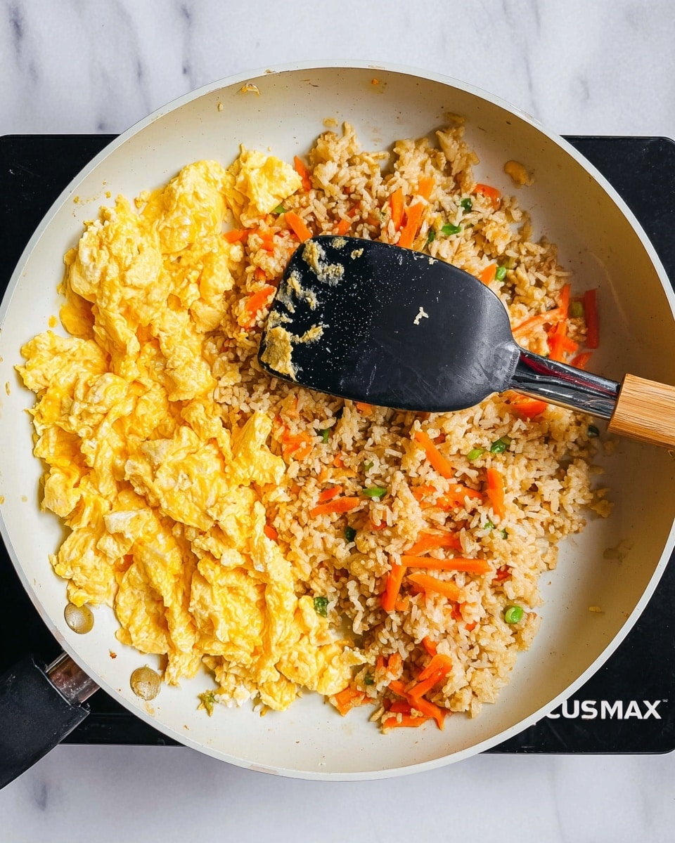 The image shows a white pan with two main parts visible: on the left side, there is a layer of soft, yellow scrambled eggs with a slightly wet texture, and on the right side, a layer of cooked fried rice mixed with small pieces of orange carrot strips and bits of green vegetables, giving it a lightly colorful look. Placed on the eggs is a black spatula with a shiny handle, and the pan sits on a black stovetop. The background surface is white marble with light veins. photo taken with an iphone --ar 4:5 --v 7
