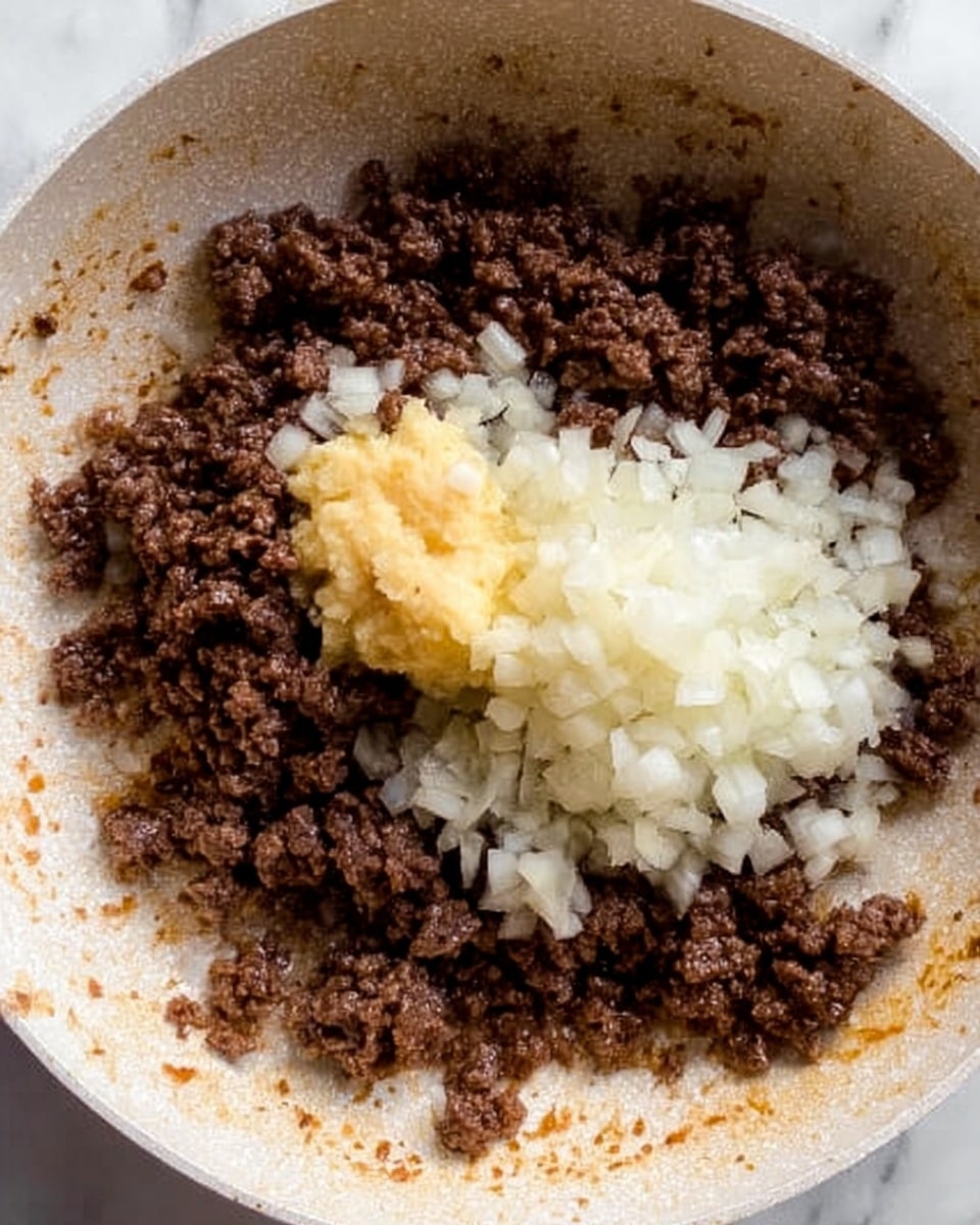 The image shows a close-up of a white pan with cooked ground beef scattered around the edges in a dark brown, crumbly texture. In the center, there is a pile of finely chopped white onions covering part of the beef. On top of the onions, there is a dollop of a light yellow, smooth paste, likely garlic or ginger, adding a soft texture contrast. The pan's bottom has browned spots from cooking, all placed on a white marbled surface. photo taken with an iphone --ar 4:5 --v 7