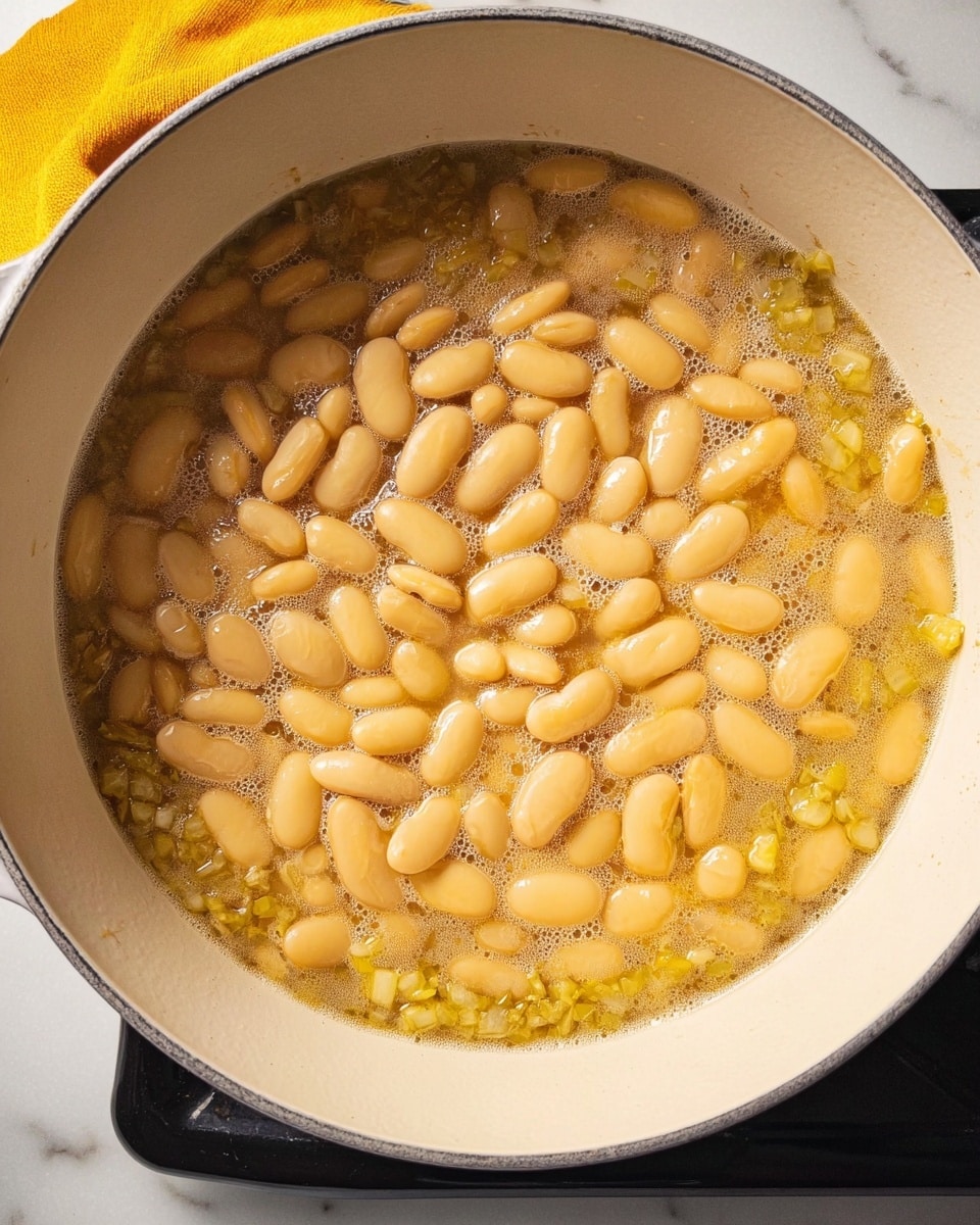 The image shows a round white pot filled with cooked yellowish beans in light broth. The beans cover most of the pot and are shiny and smooth, floating just under the surface with small bubbles scattered on top. The broth is somewhat oily with small chopped pieces of green and light brown ingredients mixed unevenly around the beans, especially near the edges. The pot rests on a white marbled surface, and the edge of a black stove and a yellow cloth can be seen at the top of the image. Photo taken with an iphone --ar 4:5 --v 7