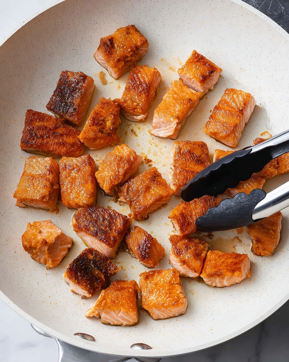 The image shows several pieces of golden brown, cooked salmon cut into small chunks, frying in a white pan with light speckled texture. The salmon pieces have a mix of crispy, browned edges and tender, slightly shiny sides with an orange-pink color. A black and silver pair of tongs is holding one piece of salmon near the bottom right of the pan. The background visible around the pan is a white marbled surface. photo taken with an iphone --ar 4:5 --v 7