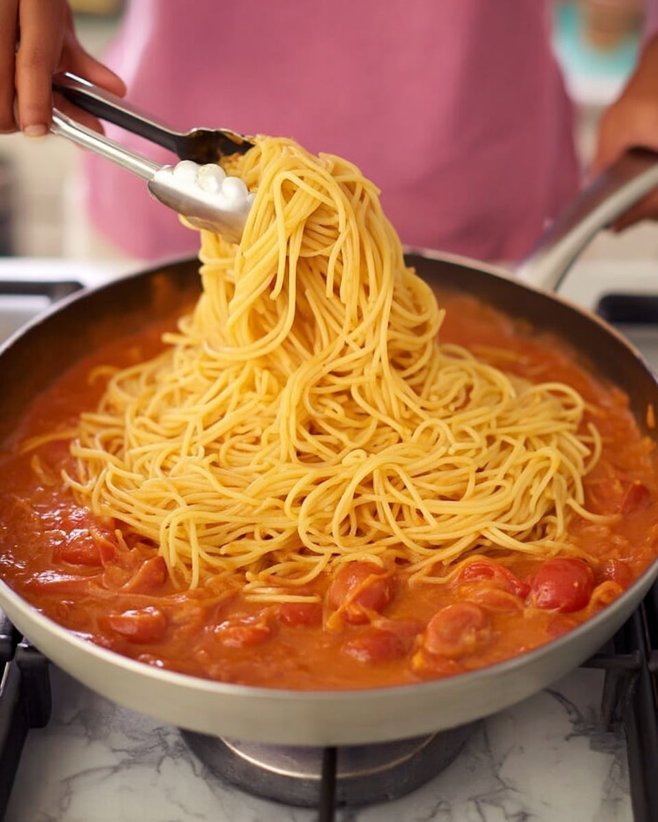The image shows a pan on a stove with a thick orange-red sauce filled with visible chunks of onions and cherry tomatoes as the base layer. On top of this sauce is a large pile of yellow cooked spaghetti noodles being added using silver tongs held by a woman's hand. The background is softly blurred, showing a person in a pink shirt holding the pan. The surface under the stove is changed to a white marbled texture. Photo taken with an iphone --ar 4:5 --v 7