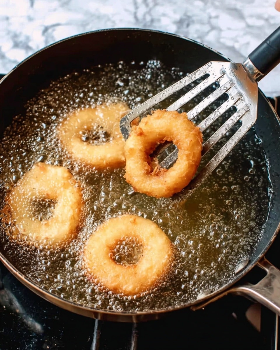 A black pan filled with bubbling hot oil is shown frying light golden brown rings of batter-coated food. A metal spatula with a black handle lifts three crispy, round fried pieces with a slightly rough texture from the oil. The background around the pan is a white marbled texture. Photo taken with an iphone --ar 4:5 --v 7