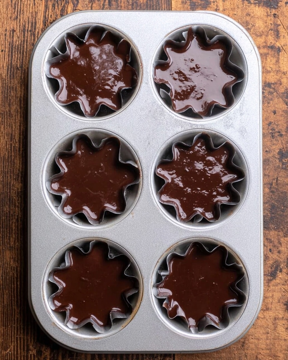 There is a silver metal baking tray with six round molds, each filled with dark brown chocolate batter. The molds have different fluted shapes, like stars and flowers, and the batter fills each mold evenly with a smooth and slightly shiny texture. The tray rests on a wooden surface that has a rough, natural wood grain. photo taken with an iphone --ar 4:5 --v 7