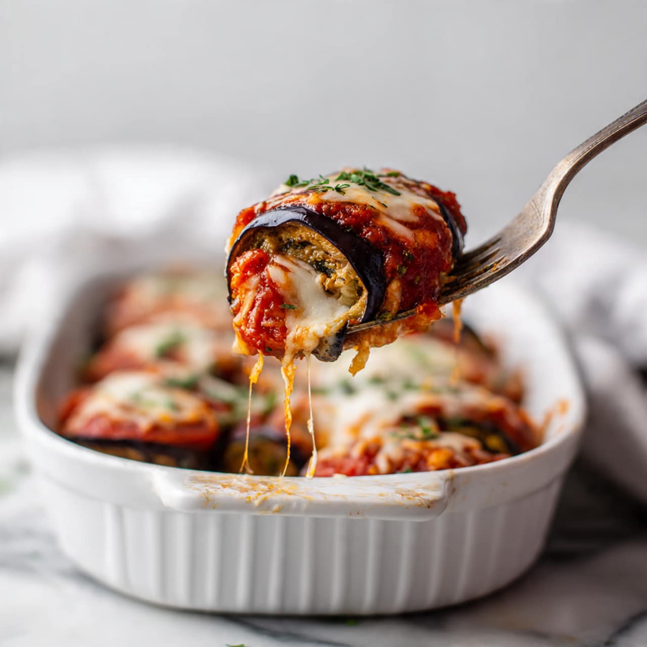 A piece of baked layered eggplant roll is held by a fork above a white ceramic baking dish filled with more of the same dish. The roll shows three main layers: the dark purple eggplant outer layer, a creamy white filling inside, and a rich red tomato sauce topped with melted golden and slightly browned cheese on top. Specks of green herbs are scattered on the cheese and sauce. The baking dish sits on a white marbled surface with soft light highlighting the textures and colors. photo taken with an iphone --ar 4:5 --v 7