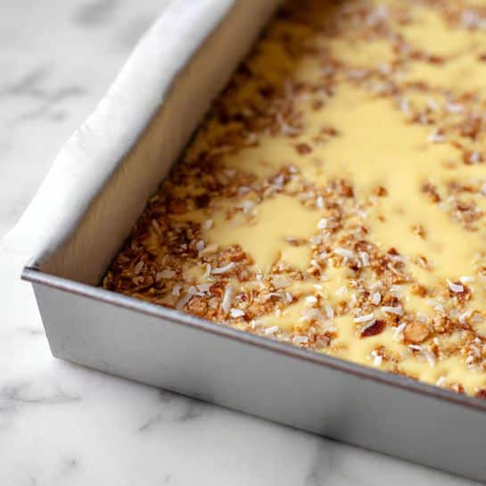 The image shows a close-up of a baking pan filled with a single thick layer of golden-brown batter. On top, there are two kinds of chocolate chips generously scattered all over the surface, one dark brown and the other light tan in color. The chips vary in size and shape but are evenly spread, creating a dotted texture against the smooth batter. The pan sits on a white marbled surface, with the focus sharply resting on the chip-covered layer, capturing the rich contrast and textures clearly. Photo taken with an iphone --ar 4:5 --v 7