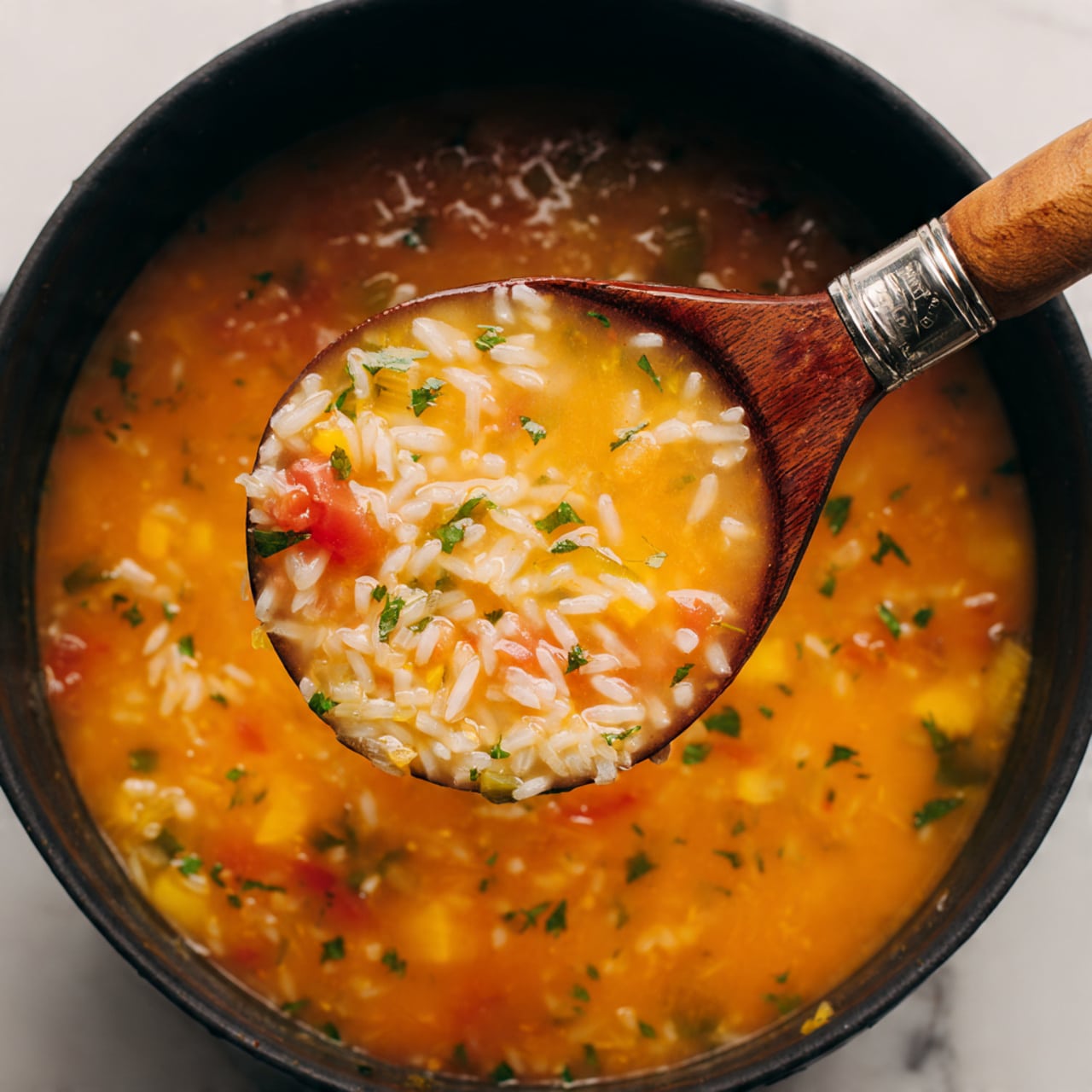 A close-up view shows a deep spoon full of chunky soup above a black pot filled with the same soup on a white marbled surface. The soup is thick and orange in color with visible cooked white rice grains, small smooth red tomato pieces, and soft yellow-orange vegetable chunks. The texture looks soft and slightly watery with mixed rice and vegetable pieces throughout. The spoon handle has a wooden grip with metal, contrasting the vibrant soup colors. photo taken with an iphone --ar 4:5 --v 7