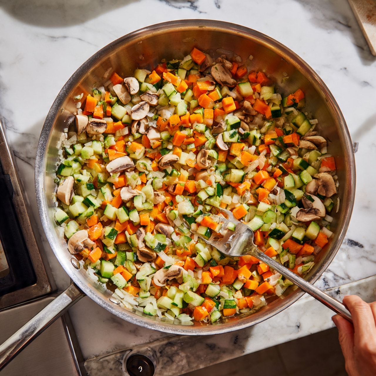 A large silver frying pan is placed on a silver stove with visible knobs at the front. Inside the pan, there is a colorful mix of chopped vegetables creating layers by size and color: small light orange carrot cubes, medium green zucchini pieces with white insides, white onion chunks, and small light brown mushroom slices. A silver spatula is stirring the vegetables, held by a visible woman's hand on the right side of the image. The background shows a white marbled counter with a faint but visible pattern. photo taken with an iphone --ar 4:5 --v 7