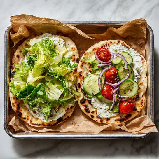 The image shows two white flatbreads placed separately on parchment paper inside a metal tray over a white marbled surface. The flatbread on the left has a layer of green leafy lettuce with a creamy white spread beneath it, creating a fresh and soft texture. The one on the right has the same base of lettuce and creamy spread but is topped with additional layers including sliced green cucumbers, halved red cherry tomatoes, and thin purple onion rings layered evenly over the lettuce. Both flatbreads have light brown char marks giving a warm, toasted appearance. The parchment paper is crumpled around the flatbreads, and the whole scene is brightly lit. Photo taken with an iphone --ar 4:5 --v 7
