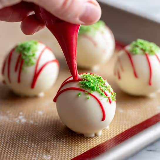 The image shows four round white chocolate balls arranged on a silicone baking mat with a golden-brown texture sitting in a tray. Each ball has two green sprinkle patches on top, giving a touch of color and texture. A woman's hand is squeezing red icing from a red piping bag to make thin red lines drizzling over the closest white chocolate ball. The surrounding surface has a white marbled texture. The focus is close up on the balls and the decoration process, showing the smooth, glossy white surface contrasted with the vibrant red icing lines and small green sprinkles. photo taken with an iphone --ar 4:5 --v 7