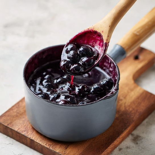 A close-up of a small gray pot filled with dark purple blueberry sauce, with whole blueberries visible in the thick liquid. Inside the pot is a gray ladle with a wooden handle, lifting a scoop of the sauce showing its syrupy texture and glossy finish. The pot is resting on a wooden cutting board on top of a white marbled textured surface beneath. Photo taken with an iphone --ar 4:5 --v 7