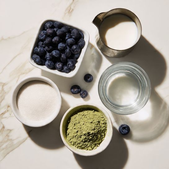 The image shows five containers of ingredients arranged on a white marbled surface. Starting from the top left, there is a small white square bowl filled with fresh dark blue blueberries. To the right of it, a small metal pitcher holds a white liquid, likely milk. Below the pitcher is a white round bowl containing clear water. On the bottom left, a white round bowl holds white granulated sugar. Finally, in the bottom center, there is a small white round bowl with a mound of green powder, possibly matcha. Two loose blueberries are placed between the bowls on the surface. The photo was taken with an iphone --ar 4:5 --v 7