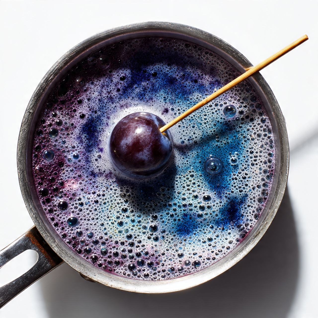 A top view of a small metal pan filled with bubbling dark purple liquid mixed with light blue and white frothy foam, with a single dark purple grape pierced on a wooden skewer resting in the middle. The pan is placed on a white marbled surface. Photo taken with an iphone --ar 4:5 --v 7