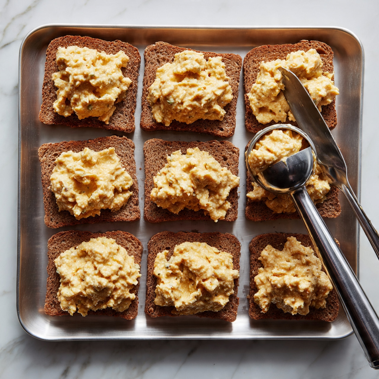 A silver tray holds multiple square slices of brown bread arranged in rows. On each slice, there is a chunky, creamy mixture with a yellowish tint, being scooped and placed using a shiny metal ice cream scooper. The bread slices are evenly spaced on the tray, which rests on a white marbled surface. Photo taken with an iphone --ar 4:5 --v 7
