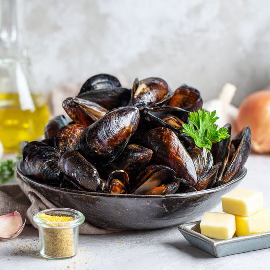A dark bowl full of shiny brown and black mussels, piled high to show many layers of shells with a small green parsley leaf on top to add color. The bowl is placed on a white marbled surface with a small gray dish holding two pale butter squares and a small jar filled with a yellow spice next to it. Around the bowl are some garlic, a glass bottle with olive oil, and some onions blurred in the background, giving a natural and fresh feeling. The lighting is soft, showing the shine on the shells clearly. photo taken with an iphone --ar 4:5 --v 7