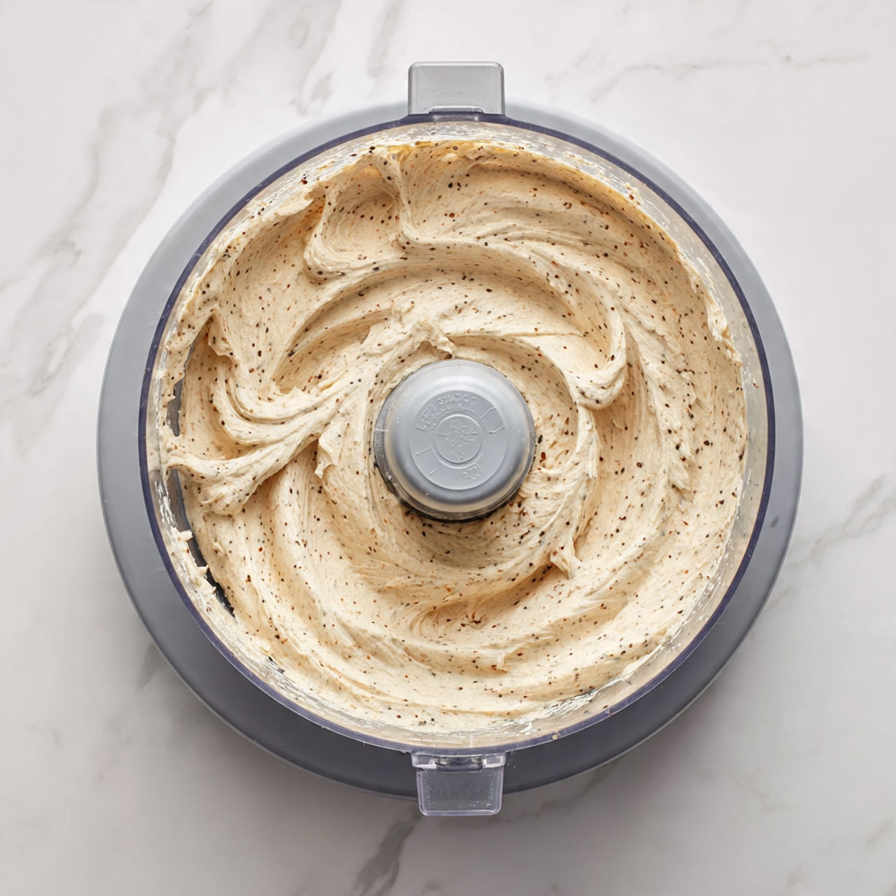 A clear plastic food processor bowl filled with a creamy mixture that has a light beige color with visible small dark specks, showing a thick and textured consistency. The mixture is swirled unevenly inside the bowl with a raised edge near the sides. The base of the food processor is gray and the whole setup is placed on a white marbled surface. photo taken with an iphone --ar 4:5 --v 7