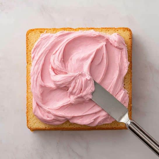 A square plain cake layer sits on a white marbled surface, topped with one thick layer of smooth, bright pink frosting being spread evenly with a metal spatula. The cake's edges are golden brown and the inside is a soft, light beige color. The frosting has a creamy texture with gentle swirls and peaks forming as it is spread across the center of the cake. Photo taken with an iphone --ar 4:5 --v 7
