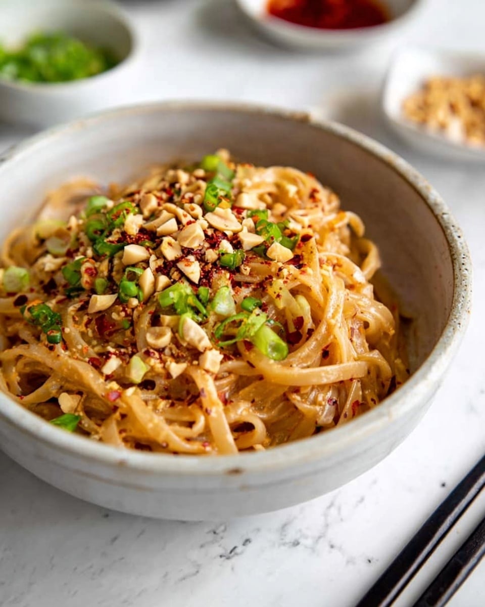A white bowl filled with creamy noodles showing a light brown sauce coating each strand. The noodles are topped with bright green sliced scallions and roughly chopped light tan peanuts. There is a sprinkle of red chili flakes adding small red specks over the dish. Black chopsticks rest on the edge of the bowl. In the background, there are blurred white bowls containing red and green sauces, set on a white marbled surface. Photo taken with an iphone --ar 4:5 --v 7